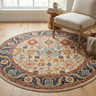 A round area rug with an intricate traditional pattern in beige, blue, red, and orange tones, styled in a bright room next to a light-colored armchair and a wooden side table.