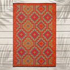 A rectangular woven rug with a Southwestern geometric pattern in red, orange, purple, and beige, laid flat on a light gray wood plank background.