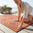 A woman kneels on a beach deck placing a rectangular outdoor rug with a geometric pattern in orange, red, purple, and beige. The background shows a sunny beach scene with potted plants and the ocean.