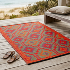 A rectangular outdoor area rug with a colorful geometric pattern in red, orange, purple, and green, laid out on a wooden deck next to a bench and sandals, with a beach background visible.