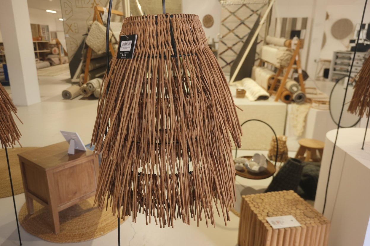 A woven wicker pendant light shade with long, thin brown strands hanging in a tapered conical shape, shown hanging indoors with a small product tag attached.