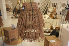 A woven wicker pendant light shade with long, thin brown strands hanging in a tapered conical shape, shown hanging indoors with a small product tag attached.