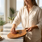 A woman in a cream linen shirt holds a round wooden serving board with a handle on a bright kitchen counter.