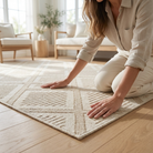 A woman kneels on a light beige woven area rug with a geometric pattern, placed on a light wood floor in a bright, modern living room.