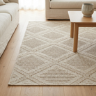 A rectangular area rug with a textured geometric diamond pattern in beige and light brown, styled on a light wood floor in a living room setting with a sofa and coffee table.
