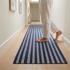 A person wearing light beige linen pants walks barefoot on a navy blue runner rug with white stripes in a bright, modern hallway with light wood floors.