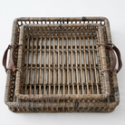 A rectangular woven rattan tray with a natural brown and beige textured body and dark brown leather handles on the sides, photographed from a top-down angle against a plain white background.