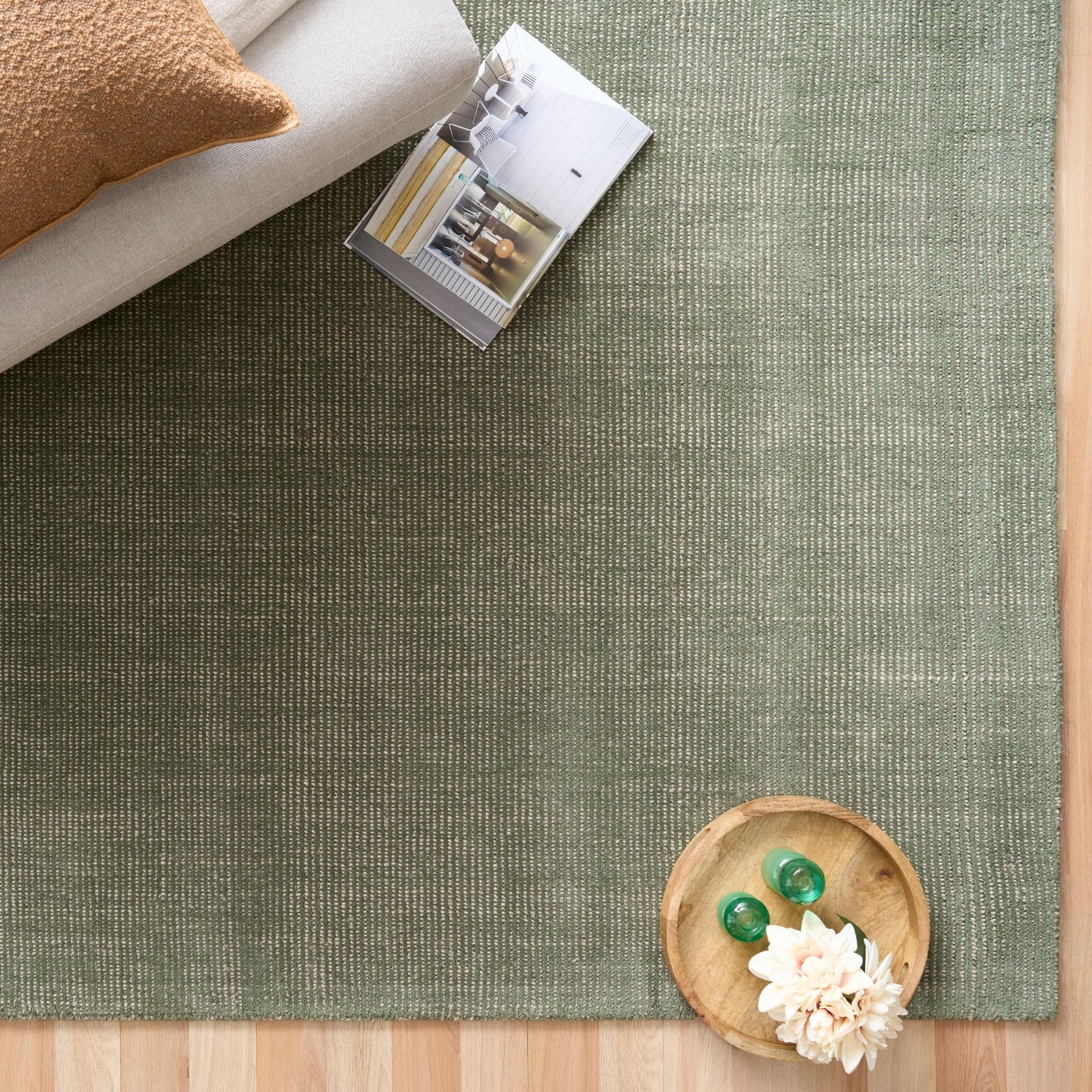 An olive green textured area rug styled in a modern living room setting on a light wood floor, with a cream sofa, a brown pillow, a magazine, and a small wooden tray with green marbles and a white flower.