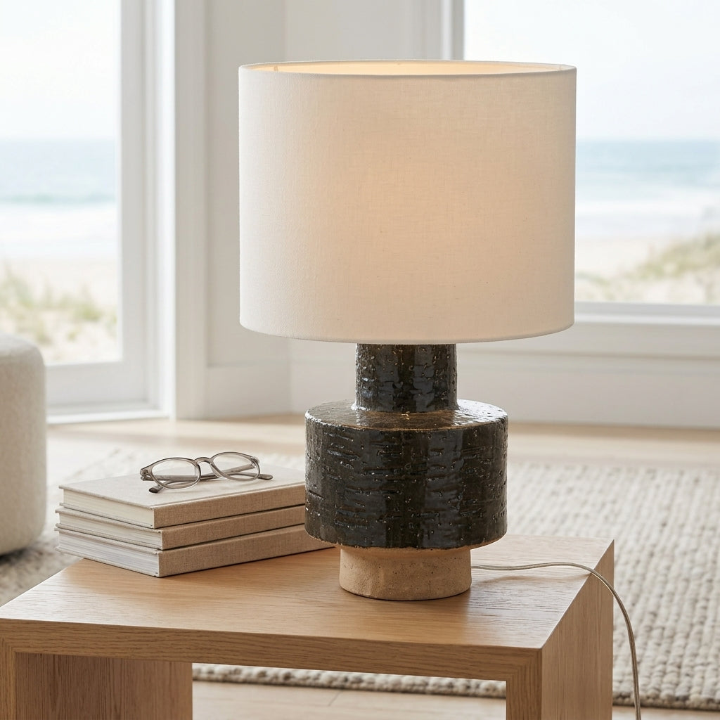 A table lamp with a warm white fabric drum shade and a textured dark gray and beige ceramic base, styled on a light wood side table in a bright room with a window showing a beach view.