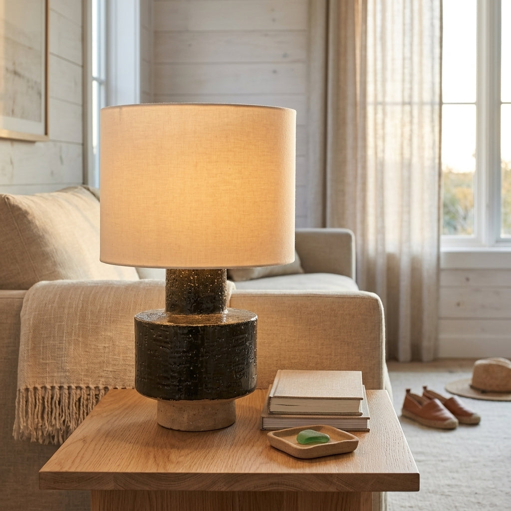 A table lamp with a warm beige fabric drum shade and a dark textured cylindrical base sits on a wooden side table in a softly lit living room. A sofa, curtains, and a hat are visible in the background.