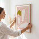 A woman in a white shirt hangs a framed minimalist art print on a white wall. The print features a light pink background with abstract white leaf shapes and a green tulip bud, enclosed in a light natural wood frame.