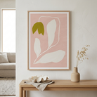 A framed minimalist botanical art print with a dusty pink background, white leaf outlines, and a single olive-green leaf, displayed on a white wall above a wooden console table with a vase and shell.