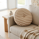 A round tufted throw pillow with beige and cream horizontal stripes rests on a light beige sofa in a bright, modern living room setting.