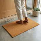 Person stepping onto a decorative doormat with a black border and brown mat, in front of a green door.
