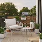 A modern outdoor patio scene with a white woven armchair, white cushion, small round white side table, beige rug, potted plants, and a wooden slat railing with string lights in the background.