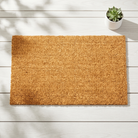 A rectangular natural coir doormat in a warm tan-brown color, laid flat on a white wooden plank floor with a small potted succulent in the top right corner.