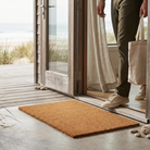 A rectangular natural coir doormat in a warm tan-brown color, positioned on a concrete floor at the threshold of a beach house. A person wearing olive green pants and beige sneakers is stepping onto the mat while holding a canvas tote bag.