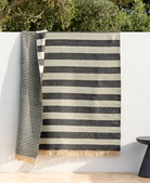 A striped woven throw blanket with cream and charcoal gray horizontal stripes and fringed edges, draped over a white wall outdoors with green foliage above.