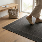 Person stepping onto a textured doormat in a home setting with a wooden bench and bag in the background.