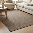 A rectangular woven area rug in a warm taupe-brown color with a subtle border, shown in a bright, modern living room setting on a light wood floor between beige sofas and a wooden coffee table.