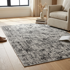 A rectangular area rug with a distressed black and gray abstract pattern, styled on a light wood floor in a bright living room next to a beige armchair.