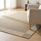 A natural woven area rug in beige and light brown tones with a checkered border, shown on a light wood floor in a bright living room next to a light-colored armchair.