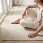 A woman in beige loungewear sits cross-legged on a textured cream-colored area rug on a light wood floor, with a window and decorative vase in the background.