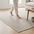 A light beige textured area rug on a light wood floor in a bright living room, with a person walking barefoot on the rug and a sofa and side table visible.