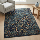 A rectangular area rug with a distressed traditional floral pattern in navy blue, beige, and muted orange, styled on a light wood floor in a living room setting next to a gray armchair and a wooden side table.