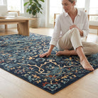A woman sits cross-legged on a large traditional-style area rug in a bright, modern living room with light wood floors and natural light coming from the windows.
