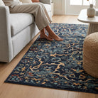A rectangular area rug with a dark navy blue background and an intricate traditional floral pattern in beige, light blue, and rust orange, shown on a light wood floor in a living room next to a sofa and a person's bare feet.
