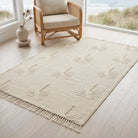 A cream-colored area rug with a raised palm leaf pattern and fringe, styled on a light wood floor next to a rattan armchair with a beach view.