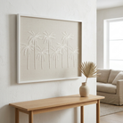 A framed wall art piece featuring white raised palm tree silhouettes on a warm beige linen-like background, displayed in a bright living room above a light wood console table with a vase.