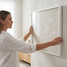 A woman in a white shirt is shown from the side, holding a white-framed wall art piece with palm tree embroidery against a textured off-white wall in a bright room.