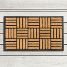A rectangular coir doormat with a black border and a woven basketweave pattern, shown flat on a light gray wood plank background.