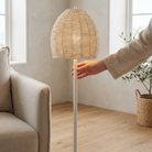 A floor lamp with a textured beige woven dome shade and a slim white pole, shown in a bright living room with a person's hand reaching toward the top. A beige armchair and a potted plant are visible in the background.
