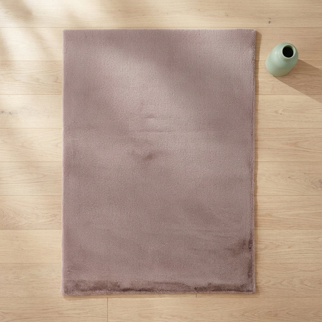 A rectangular plush rug in a muted dusty mauve color, laid flat on a light wood floor next to a small light green ceramic vase.