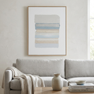 A framed abstract art print with horizontal watercolor stripes in soft beige, cream, and light blue tones, hanging on a textured off-white wall above a light gray sofa in a styled living room.