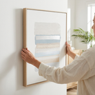 A person in a beige linen shirt holds a light wood framed abstract art print against a white wall in a bright, modern room.
