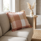 A square throw pillow with a muted plaid pattern in dusty pink, beige, and white, featuring fringed edges, resting on a light gray sofa in a bright, modern living room.