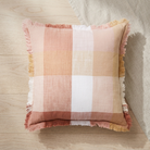 A square throw pillow with a soft plaid pattern in dusty pink, peach, cream, and white, finished with fringed edges, resting on a light wood floor next to a neutral fabric surface.