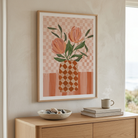 A framed art print featuring a stylized floral illustration with peach tulips in a diamond-patterned vase, hung on a beige wall above a light wood console table with a bowl, books, and a mug.