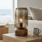 A modern table lamp with a smoky translucent glass dome and a dark wood base, lit with an exposed filament bulb, set on a wooden table in a living room with a window showing the ocean view.
