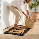 Person placing a woven bag on a doormat with a palm tree design in a bright room.