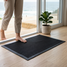 A black textured rubber door mat with a raised border, shown on a light wood floor in front of glass doors. A person's bare feet and lower legs are stepping onto the mat, and a potted green plant sits nearby.