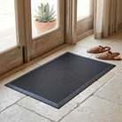 A black rectangular mat with a raised grid texture lies on a light stone floor in front of wooden glass doors, with a pair of brown sandals nearby and a potted succulent visible outside the door.