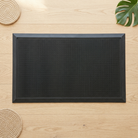 A black rectangular mat with a raised border and a dense textured surface, photographed from above on a light wood surface with woven placemats and a green leaf as styled props.