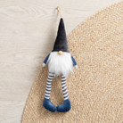 A small hanging gnome decoration with a black pointed hat, white beard, blue sleeves and shoes, and striped blue-and-white legs, photographed on a woven beige mat over a light wood surface.