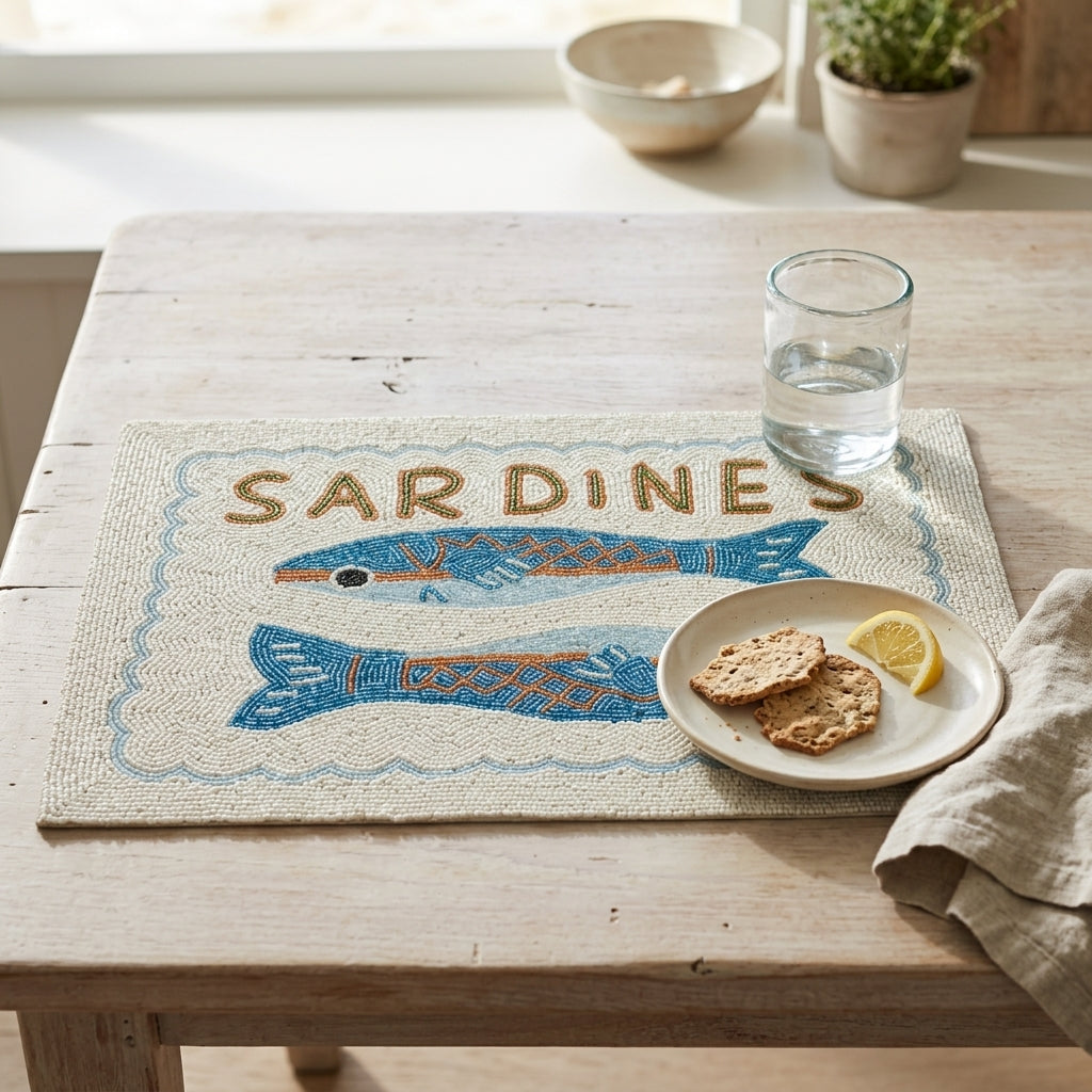 A woven rectangular placemat with a blue and orange sardine design and the word “SARDINES” in raised letters, styled on a light wood table with a glass of water, cookies on a plate, and a napkin.