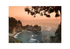 A scenic landscape photograph of a rocky coastal inlet at sunset, with orange sky, blue-green ocean water, dark cliffs, and tree silhouettes framing the top and right edges, shown on a white background.